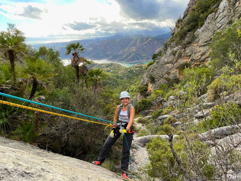 Woman rappelling down a dry wall in Monchalbán Canyon, with a breathtaking backdrop of rivers, mountains and the Mediterranean coast of Marbella.